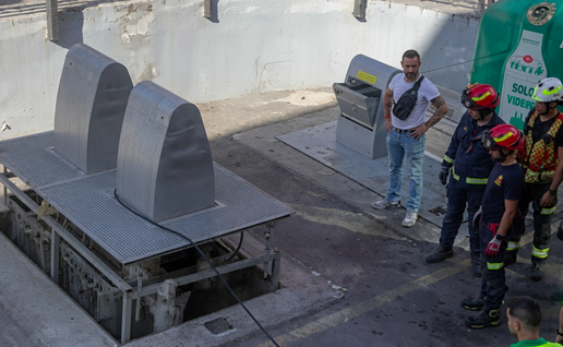 Emergency responders and firefighters gathered around a large metal underground rubbish chute on a paved outdoor site, with one civilian man in a white t-shirt observing.