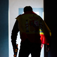 A man seen from behind, with a Spanish flag draped over his shoulder and holding a beer bottle, stumbling as he enters a dark room.