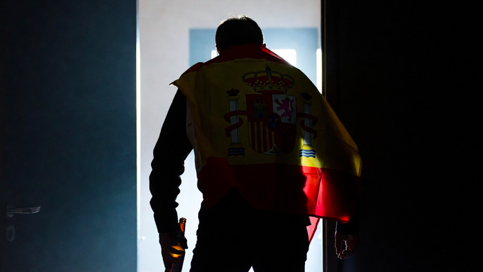 A man seen from behind, with a Spanish flag draped over his shoulder and holding a beer bottle, stumbling as he enters a dark room.