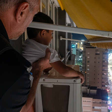A man with short gray hair in a dark shirt stands behind a young boy with short dark hair in a white t-shirt. The boy leans on a white outdoor air-conditioning unit on a high-rise balcony at night, looking out toward a city skyline and illuminated beach. A yellow awning is visible overhead, and the man’s hand rests supportively on the boy’s back.