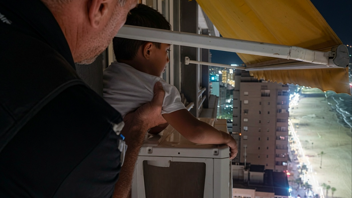 A man with short gray hair in a dark shirt stands behind a young boy with short dark hair in a white t-shirt. The boy leans on a white outdoor air-conditioning unit on a high-rise balcony at night, looking out toward a city skyline and illuminated beach. A yellow awning is visible overhead, and the man’s hand rests supportively on the boy’s back.