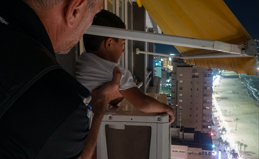 A man with short gray hair in a dark shirt stands behind a young boy with short dark hair in a white t-shirt. The boy leans on a white outdoor air-conditioning unit on a high-rise balcony at night, looking out toward a city skyline and illuminated beach. A yellow awning is visible overhead, and the man’s hand rests supportively on the boy’s back.
