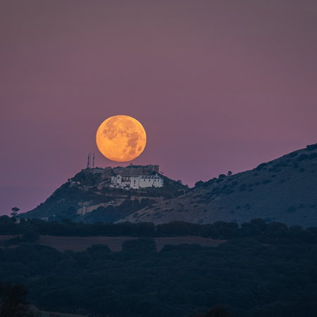A bright full moon rises above a hilltop village in Spain at dusk, with rolling countryside silhouetted beneath a pink evening sky.