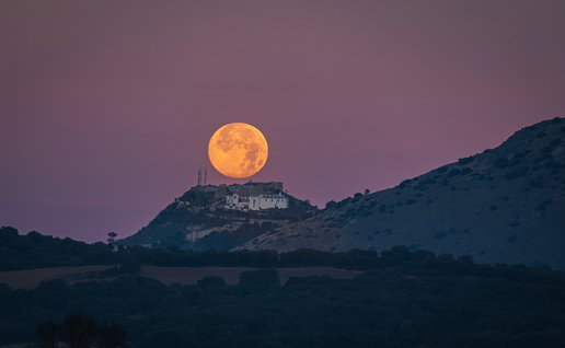 A bright full moon rises above a hilltop village in Spain at dusk, with rolling countryside silhouetted beneath a pink evening sky.