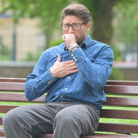 A man with glasses sitting on a park bench, holding his hand over his chest while coughing, illustrating potential heart strain or shortness of breath.