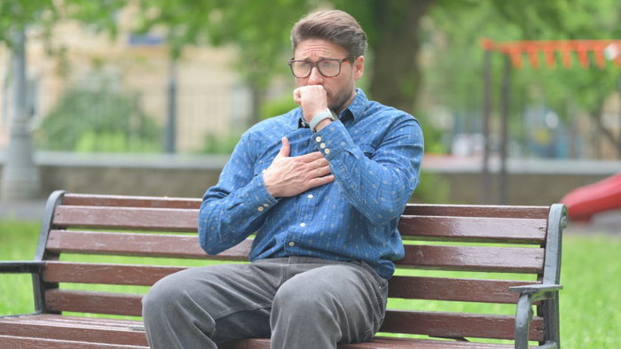 A man with glasses sitting on a park bench, holding his hand over his chest while coughing, illustrating potential heart strain or shortness of breath.