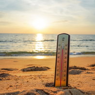 A thermometer stands on a sandy beach beside the sea, illustrating warm coastal waters during the Spanish summer.