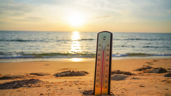 A thermometer stands on a sandy beach beside the sea, illustrating warm coastal waters during the Spanish summer.