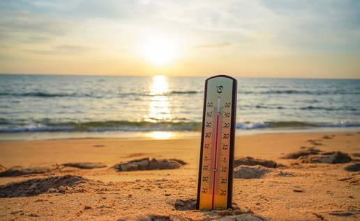 A thermometer stands on a sandy beach beside the sea, illustrating warm coastal waters during the Spanish summer.