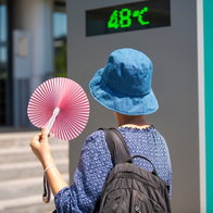 Woman wearing a hat, holding a fan andlooking at street thermometer and extreme temperature marking 48 degrees celsius on hot summer day. Extreme weather conditions.
