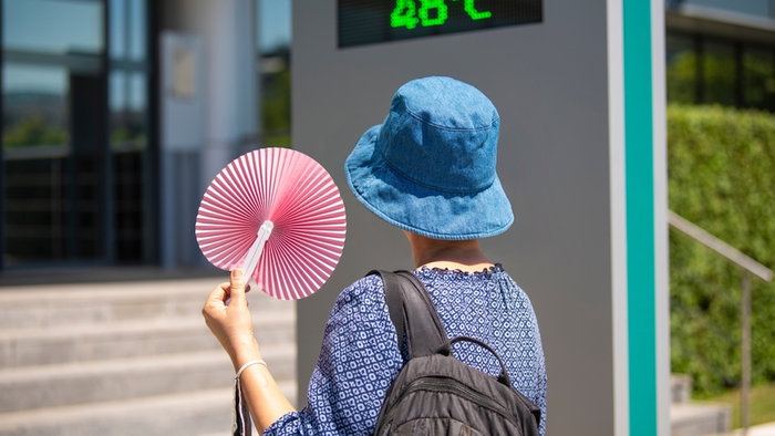 Woman wearing a hat, holding a fan andlooking at street thermometer and extreme temperature marking 48 degrees celsius on hot summer day. Extreme weather conditions.