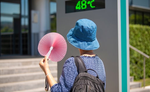 Woman wearing a hat, holding a fan andlooking at street thermometer and extreme temperature marking 48 degrees celsius on hot summer day. Extreme weather conditions.