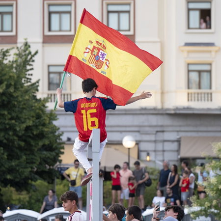 Boy holds Spanish flag whilst balancing on a metal bar waiting for the Spanish football team to arrive