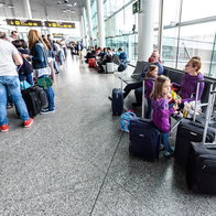 Passengers queue with suitcases inside a busy airport terminal during the summer holiday travel season.