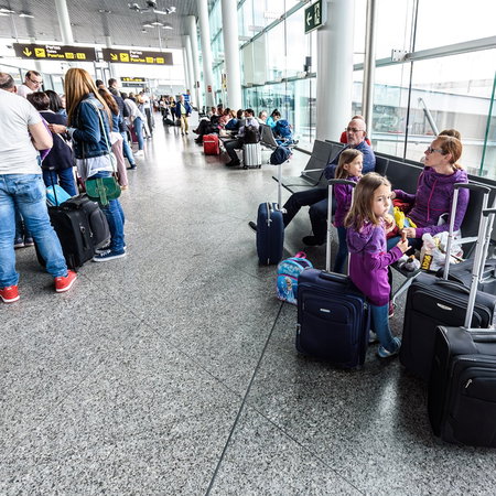 Passengers queue with suitcases inside a busy airport terminal during the summer holiday travel season.