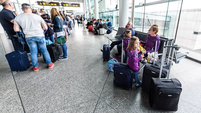 Passengers queue with suitcases inside a busy airport terminal during the summer holiday travel season.