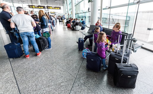 Passengers queue with suitcases inside a busy airport terminal during the summer holiday travel season.