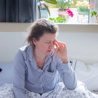 A fatigued woman in pajamas sitting on a bed rubbing her eyes, illustrating tiredness and exhaustion from potential vitamin B12 deficiency symptoms.