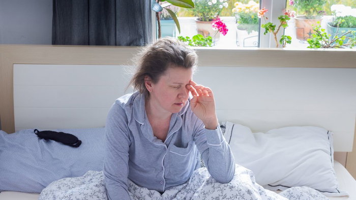 A fatigued woman in pajamas sitting on a bed rubbing her eyes, illustrating tiredness and exhaustion from potential vitamin B12 deficiency symptoms.