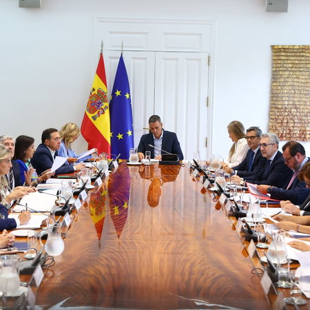 Spanish Government gathered at a table holding a cabinet meeting with the Spanish and European flags in the background