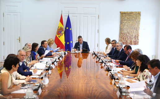 Spanish Government gathered at a table holding a cabinet meeting with the Spanish and European flags in the background