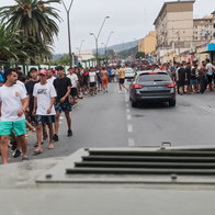 Crowd of young men lining a city street as a gray car drives away, with armored vehicle in foreground.