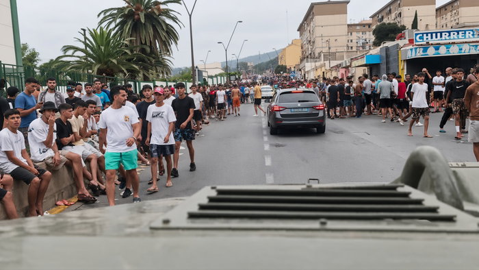 Crowd of young men lining a city street as a gray car drives away, with armored vehicle in foreground.