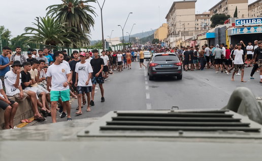 Crowd of young men lining a city street as a gray car drives away, with armored vehicle in foreground.