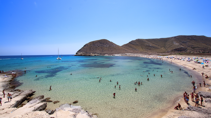 View from above of the Playazo beach in Rodalquilar, Níjar, whilst people swim in the water and sunbathe on the sand.