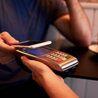 A customer taps a smartphone against a card payment terminal to pay at a café table.