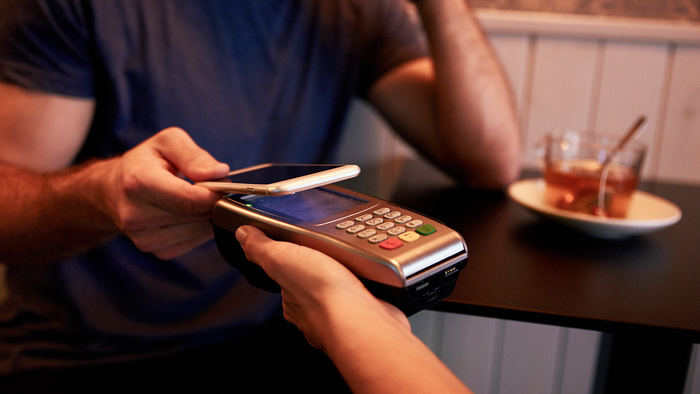 A customer taps a smartphone against a card payment terminal to pay at a café table.