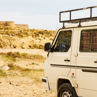 A white camper van with a roof rack parked on a dirt track near sandy coastal cliffs and a small stone fort in Spain.
