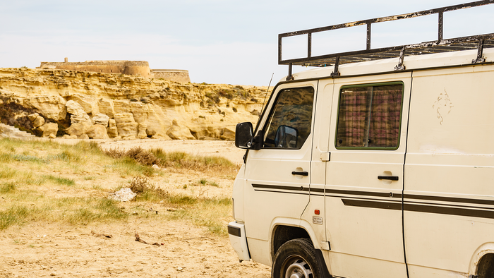 A white camper van with a roof rack parked on a dirt track near sandy coastal cliffs and a small stone fort in Spain.