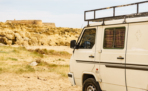 A white camper van with a roof rack parked on a dirt track near sandy coastal cliffs and a small stone fort in Spain.