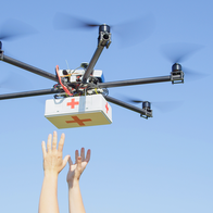 A drone carrying a first aid box flies above a pair of raised hands against a blue sky.