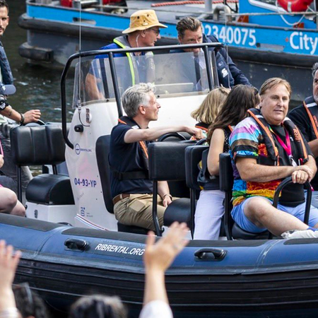 Rob Jetten in a white shirt and black life vest waving from a boat beside spectators at Amsterdam Pride.