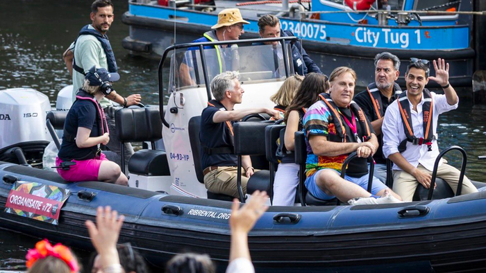 Rob Jetten in a white shirt and black life vest waving from a boat beside spectators at Amsterdam Pride.