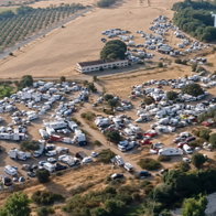 Aerial view of a large rural campsite packed with hundreds of white RVs, vans and cars near a building, dry fields, trees and a small lake, with Guardia Civil logo.