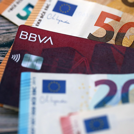 Euro banknotes in various denominations fanned out on a wooden surface, with a BBVA bank card tucked among them.