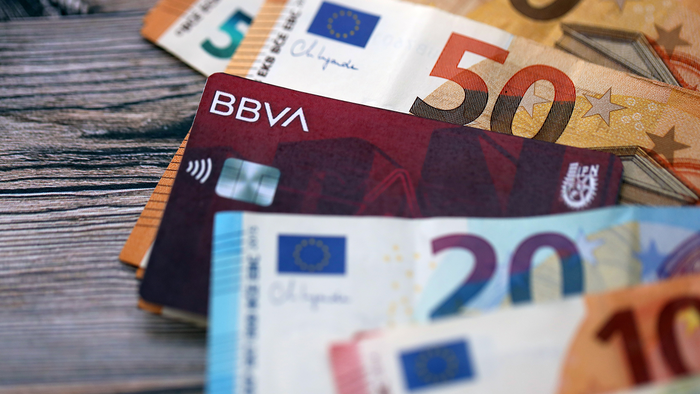 Euro banknotes in various denominations fanned out on a wooden surface, with a BBVA bank card tucked among them.