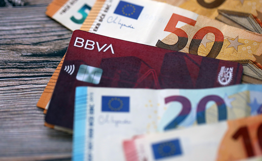 Euro banknotes in various denominations fanned out on a wooden surface, with a BBVA bank card tucked among them.