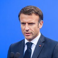 Emmanuel Macron speaking at a podium in a dark suit against a blue backdrop.