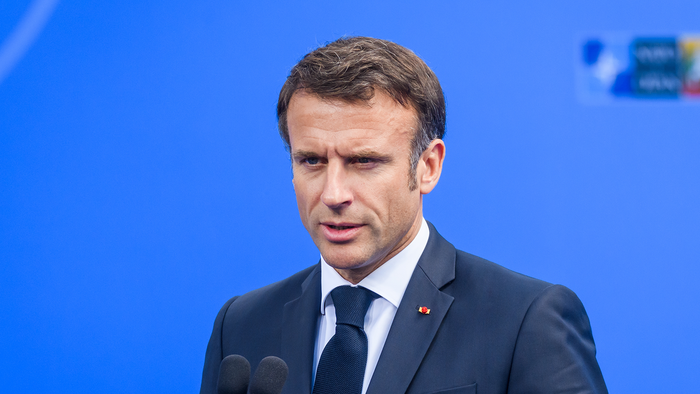 Emmanuel Macron speaking at a podium in a dark suit against a blue backdrop.