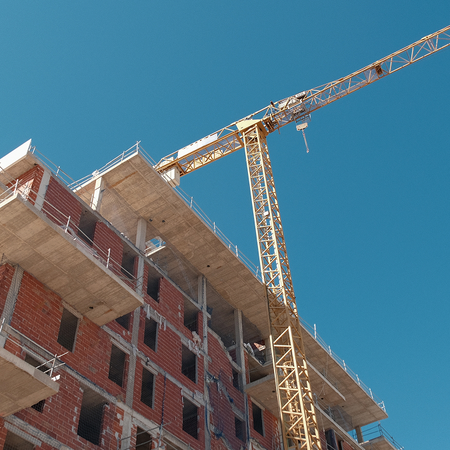 A crane towers over an apartment block under construction, with exposed brick walls and unfinished balconies against a clear blue sky.