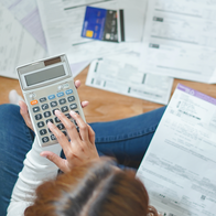 A woman sitting cross-legged on a wooden floor, using a calculator surrounded by household bills and a credit card.