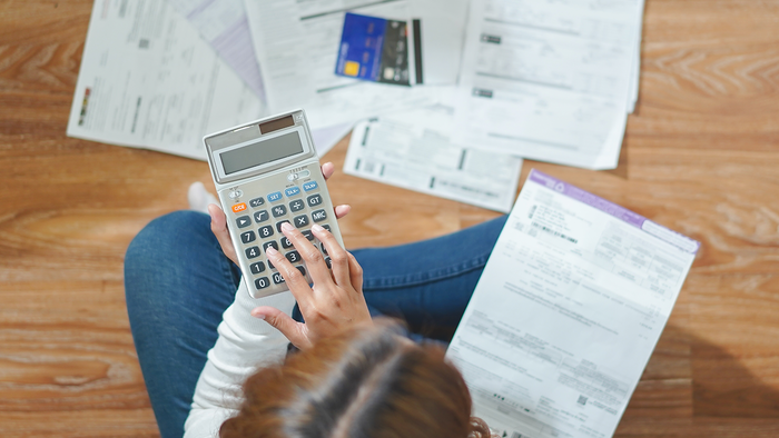 A woman sitting cross-legged on a wooden floor, using a calculator surrounded by household bills and a credit card.