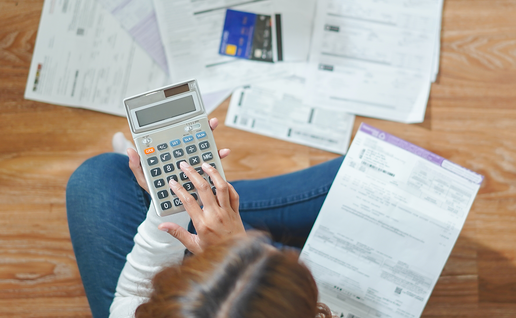 A woman sitting cross-legged on a wooden floor, using a calculator surrounded by household bills and a credit card.