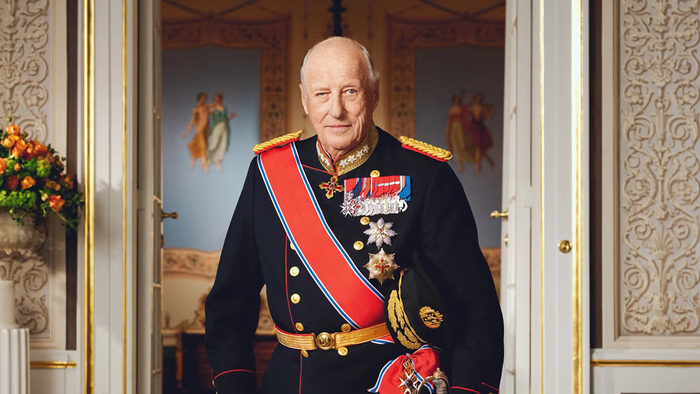 King Harald V of Norway standing in official military dress uniform with medals and sash, in a palace doorway.