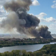 Thick black smoke rising from an industrial fire near a river, with the London skyline visible in the background.