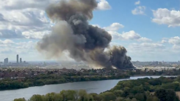 Thick black smoke rising from an industrial fire near a river, with the London skyline visible in the background.